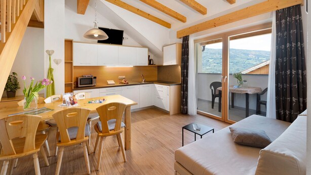Eine moderne Küche mit Holzbalken, eine Essecke mit Holzsesseln, eine Couch und Blick ins Freie.