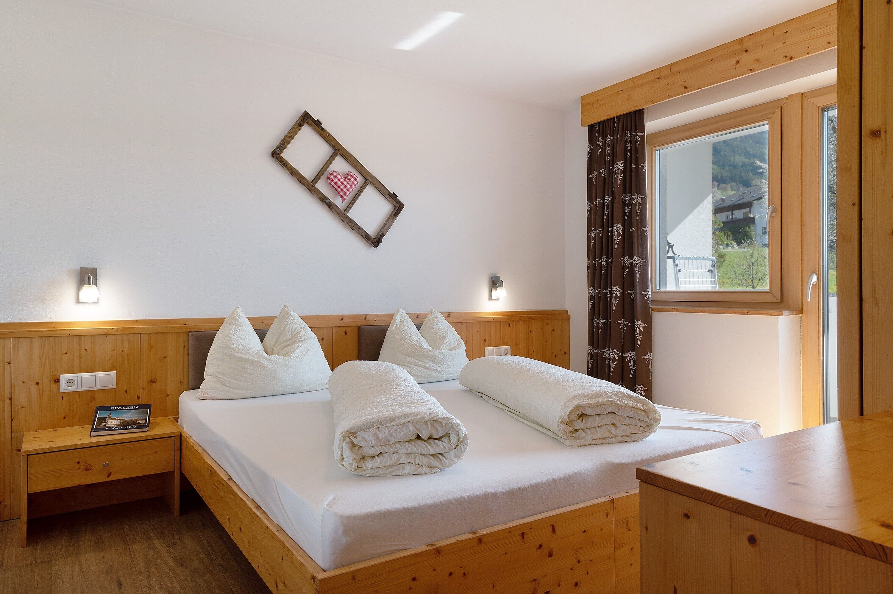 Una cama con estructura de madera, ropa de cama y almohadas blancas, una mesita de noche con un libro, una lámpara de pared y una ventana con cortinas.