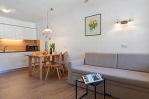 A modern kitchen with white cabinets, a dining table with chairs, a microwave, and a small plant. There is a beige sofa and a magazine on a side table.