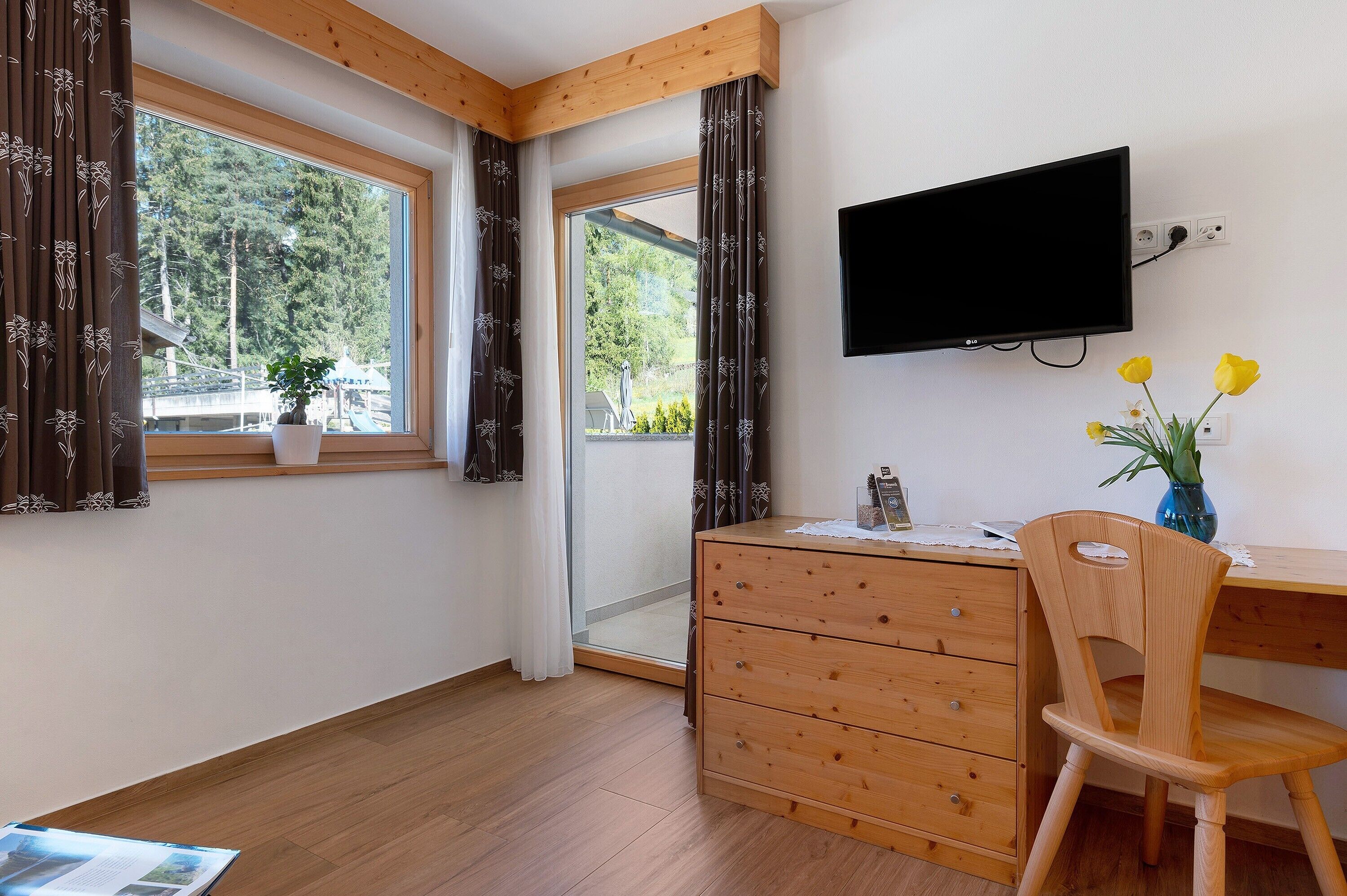 A room with a wooden desk, a chair, a TV mounted on the wall, and a window with curtains.