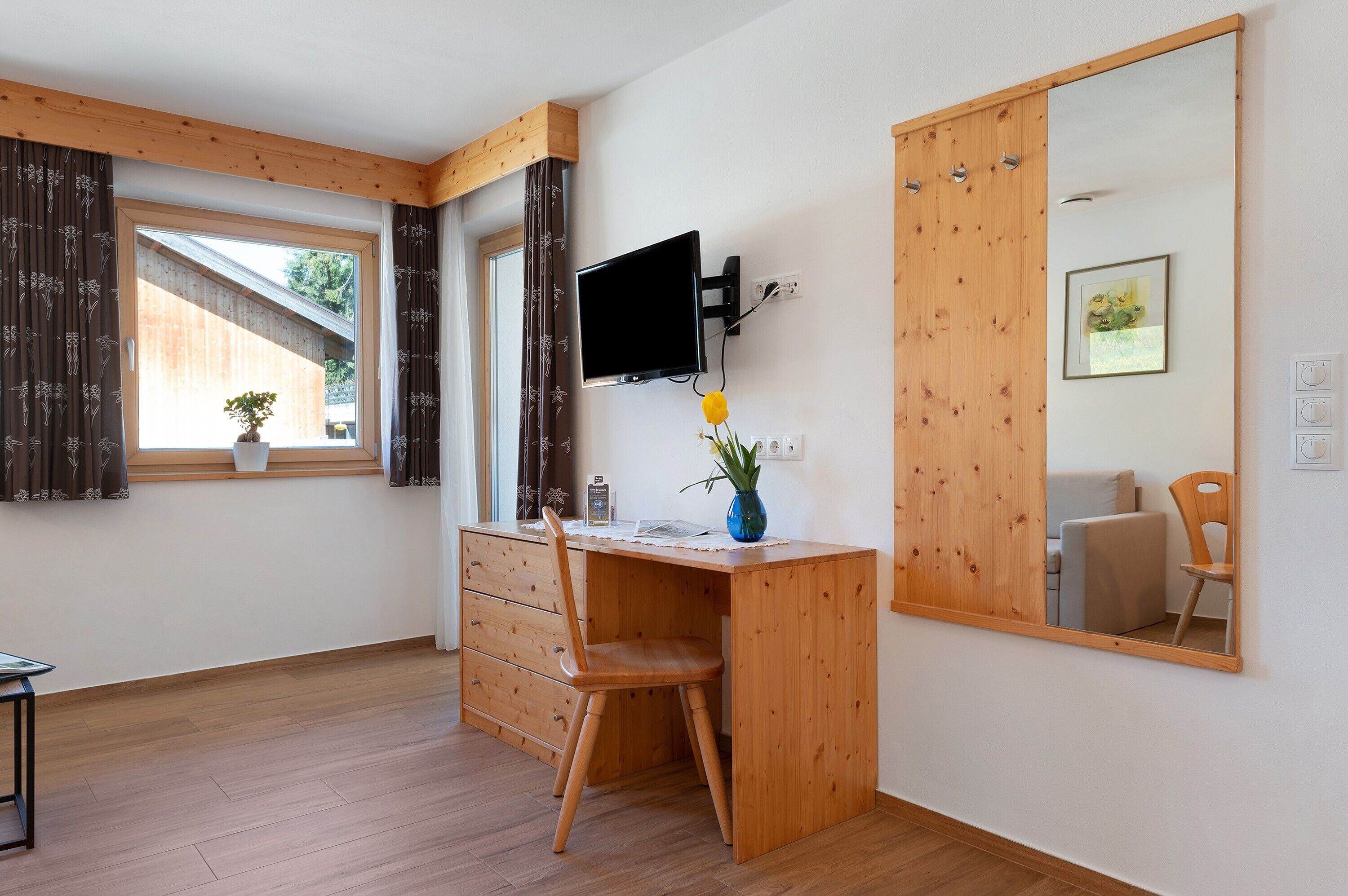 A room with a wooden desk, a chair, a TV, a mirror, a window with curtains, a potted plant, and a small table.