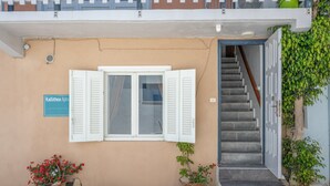 Ein Gebäude mit beige-farbener Fassade, einem Schild mit der Aufschrift 'Kallithea Apts', einem Fenster mit weißen Fensterläden, einer Treppe und Topfpflanzen.