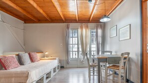 Ein Zimmer mit Bett, Tisch, Stühlen, einem Fenster mit Vorhängen und einer Holzbalkendecke.