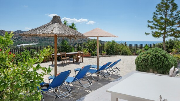 Eine Terrasse am Strand mit blauen Liegestühlen, einem Tisch und einem Strohdachschirm.