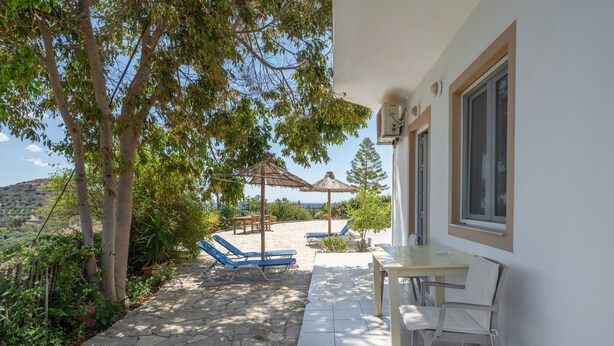 Une terrasse avec un sol en pierre, une table et des chaises, entourée d’arbres et offrant une vue sur le paysage.