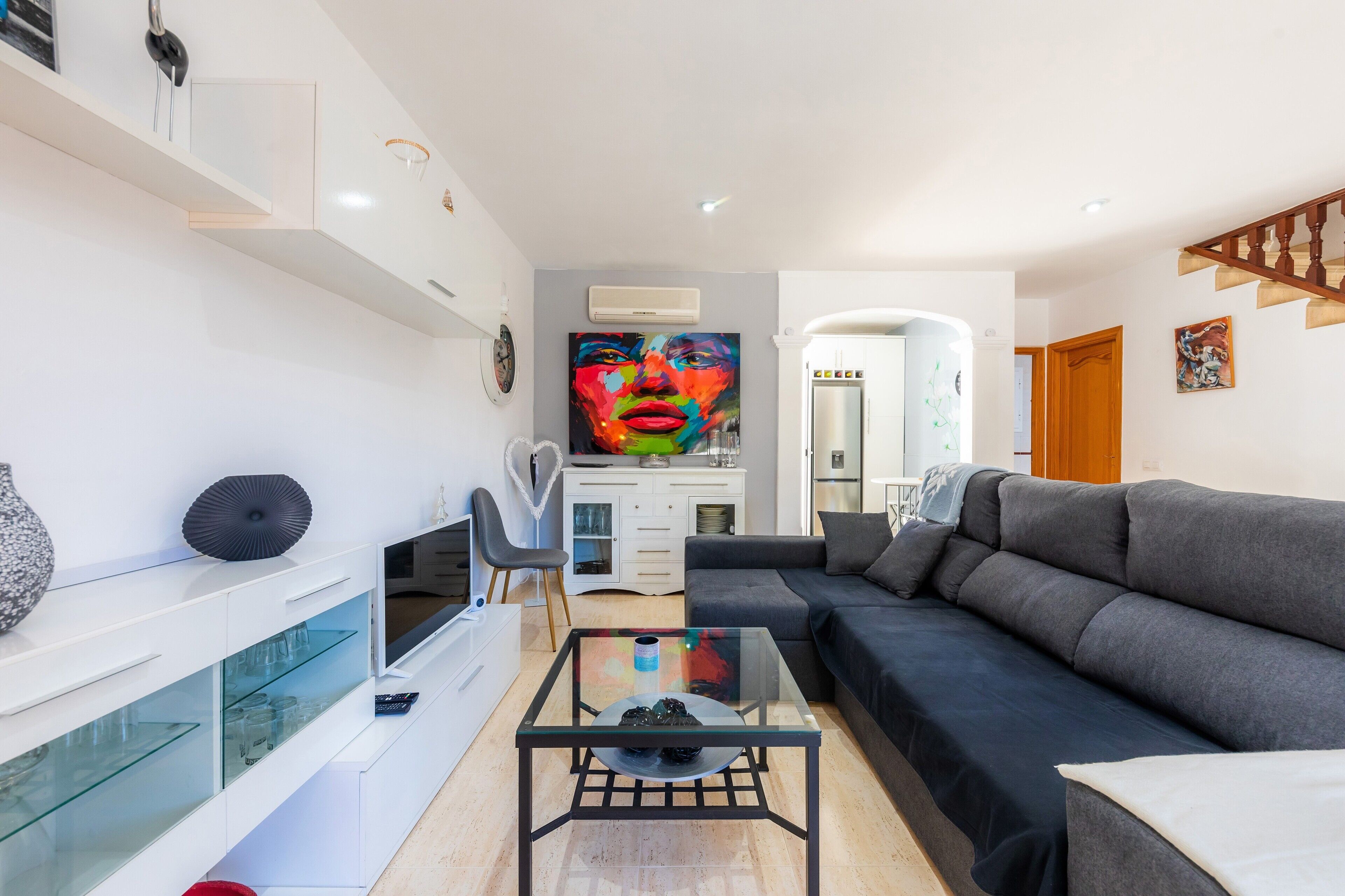 A modern living room with a glass coffee table, a black sofa, a white entertainment unit, and a large painting on the wall.