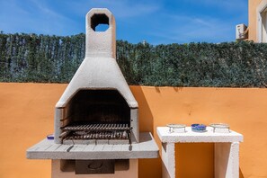 A concrete outdoor grill with a chimney, a small table with dishes, and a hedge in the background. - Holiday Home 'Eulalia Beach' with Private Terrace, Wi-Fi and Air Conditioning (Can Picafort)