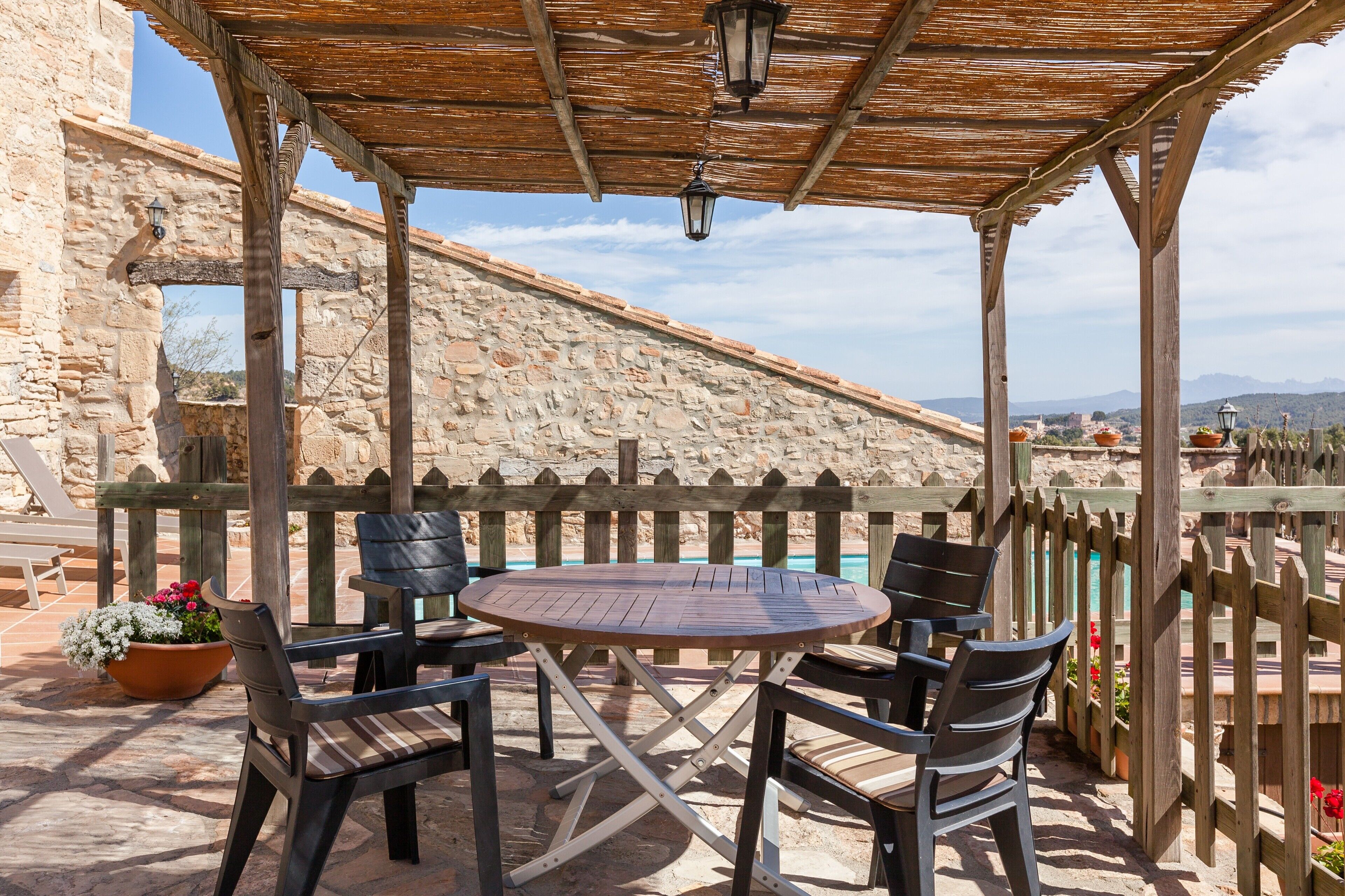 Una pérgola de madera con una mesa y sillas, paredes de piedra y vistas al paisaje.