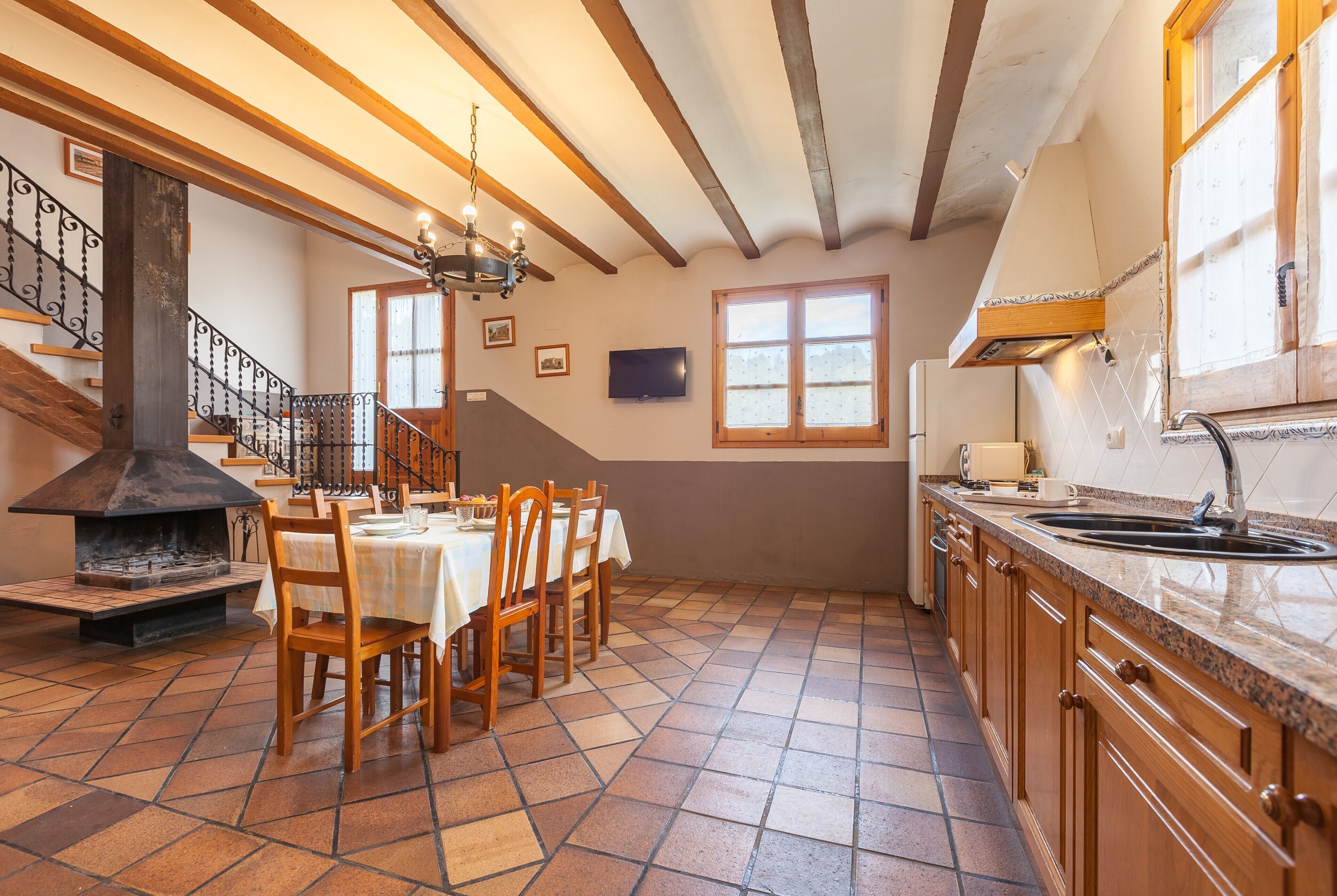 A kitchen with a tiled floor, wooden cabinets, a dining table with chairs, a fireplace, and a staircase.