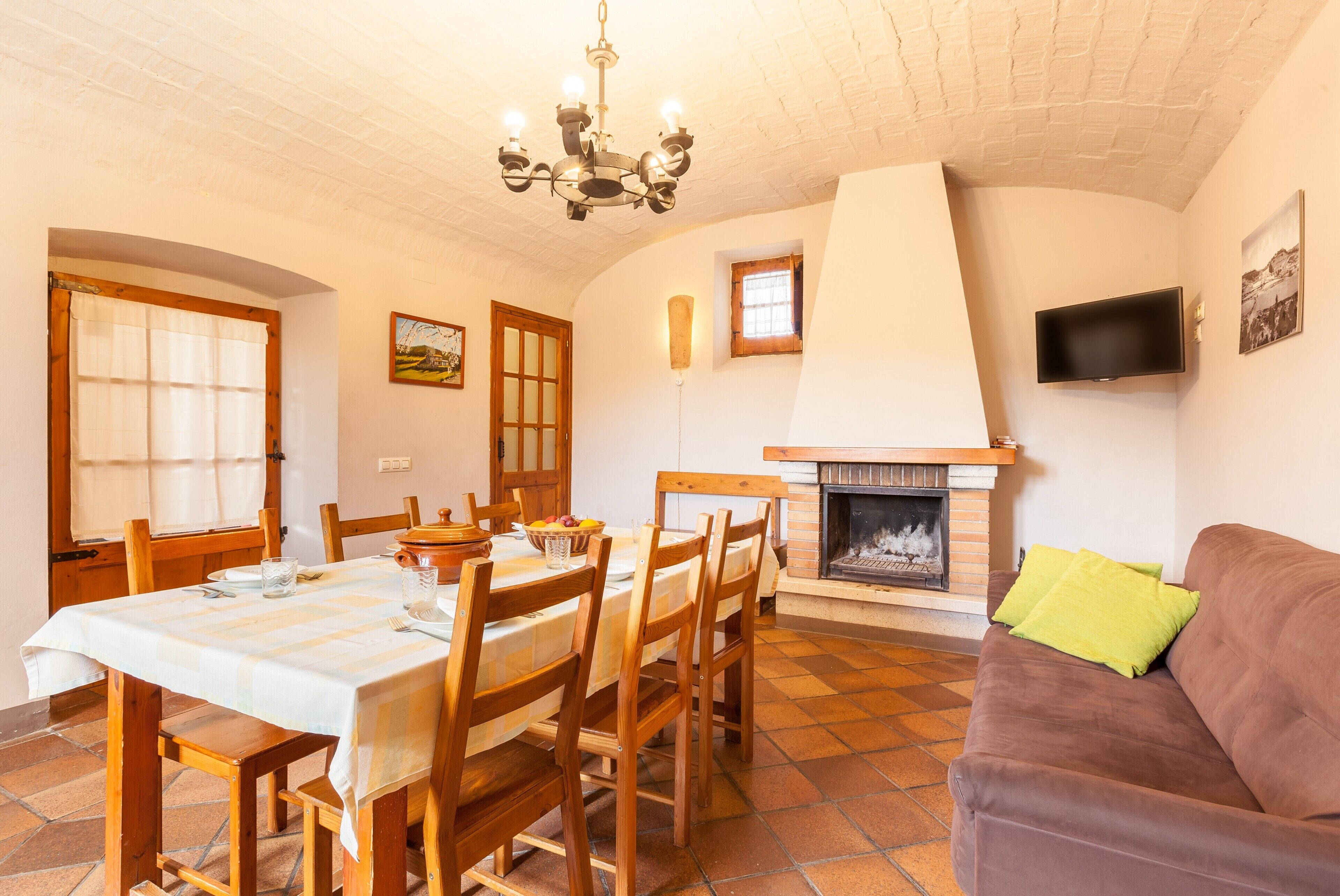Un comedor con una mesa preparada para cuatro personas, sillas de madera, una chimenea, un televisor montado en la pared y un sofá marrón.