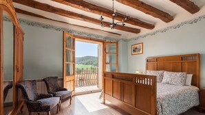 Een slaapkamer met een houten bed, twee gestoffeerde fauteuils, een houten garderobe en een groot raam met uitzicht op een groen landschap.