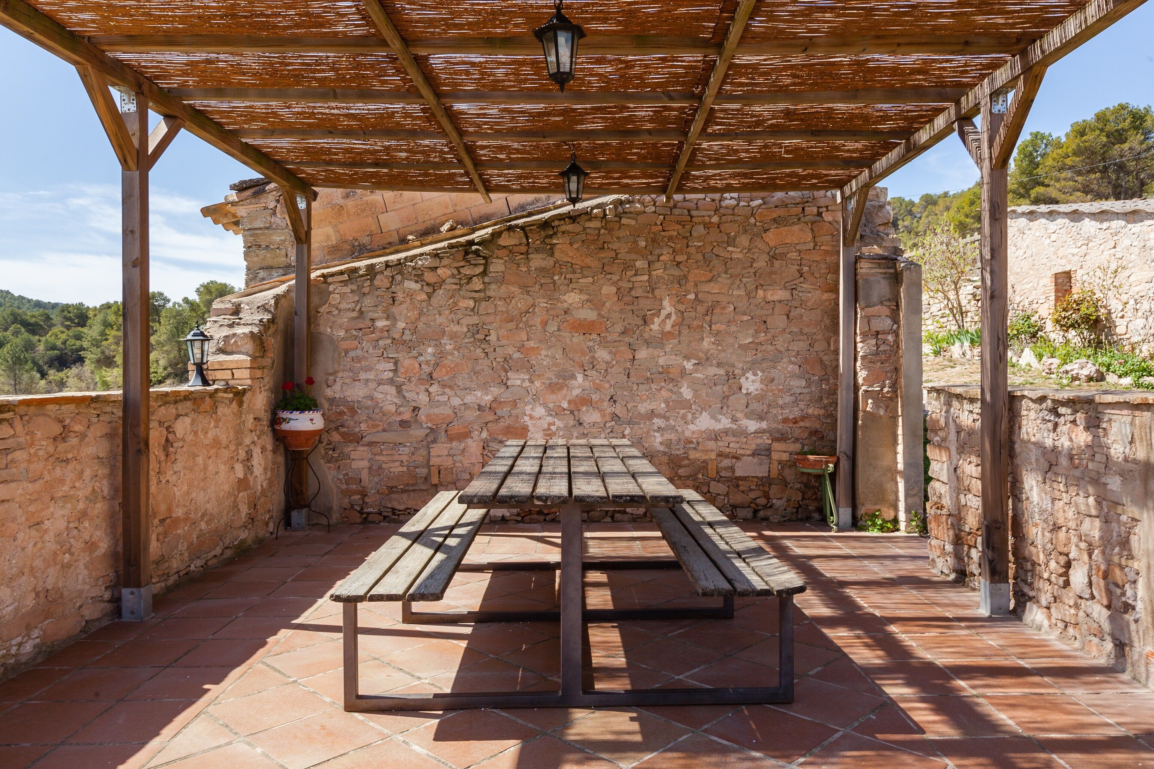 A covered outdoor area with a stone wall, a wooden table with benches, and hanging lanterns.