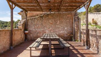 A covered outdoor area with a stone wall, a wooden table with benches, and hanging lanterns.