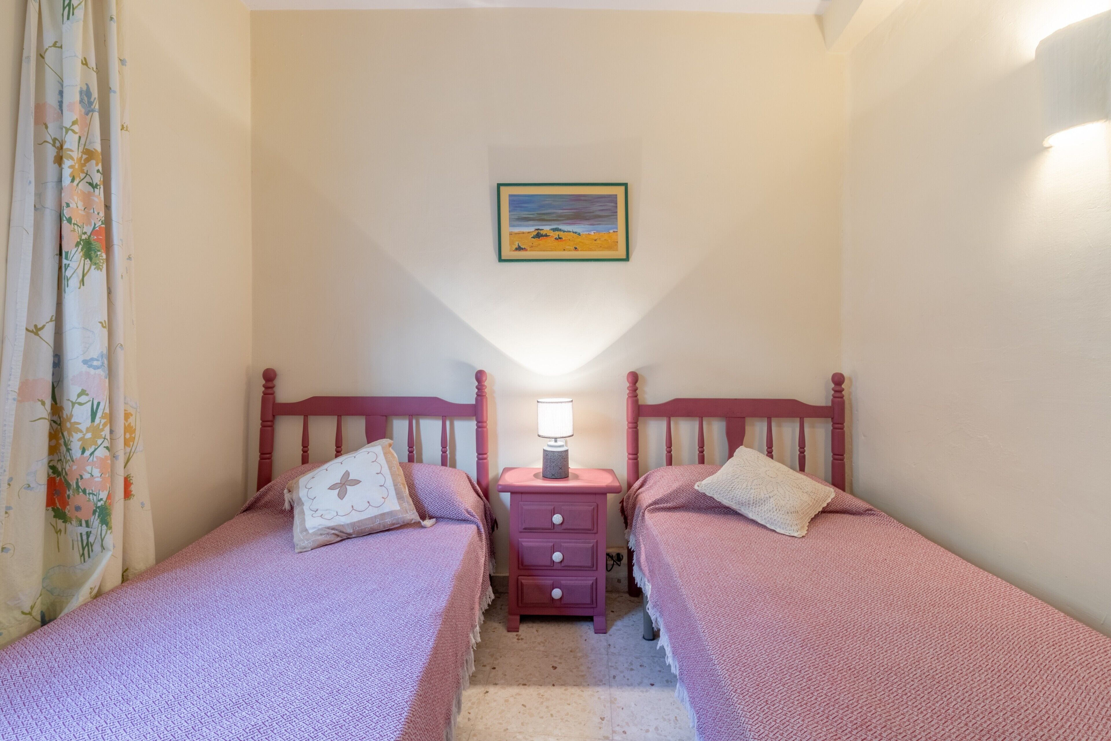 A bedroom with two beds, a nightstand with a lamp, a framed picture on the wall, and a floral curtain.