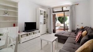 Ein Wohnzimmer mit Fernseher, weißen Möbeln, einer Couch und Blick ins Freie.