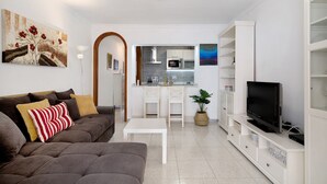 Ein Wohnzimmer mit einer Couch, einem Couchtisch, einem Fernseher auf einem Schrank, einer Küche im Hintergrund und einer weißen Schrankwand mit Regalen.