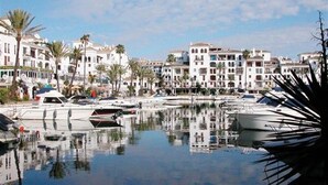 A marina with boats docked, surrounded by multi-story buildings with balconies, palm trees, and a clear sky. - Apartment 'Residencial Duquesa Fase I' with Terraces, Shared Pools & Wi-Fi (Manilva)