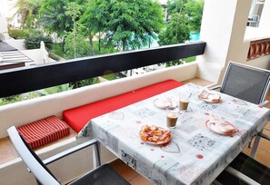 Un balcón con una mesa preparada para dos personas, un banco con cojines rojos y vistas a una piscina y vegetación.