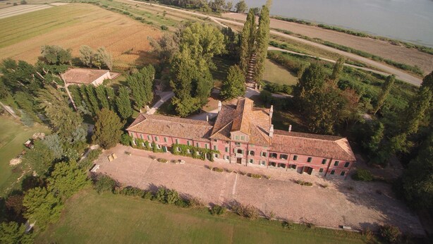 Une vue aérienne d’un grand bâtiment historique doté de plusieurs ailes, entouré de vastes champs et avec une rivière en arrière-plan.