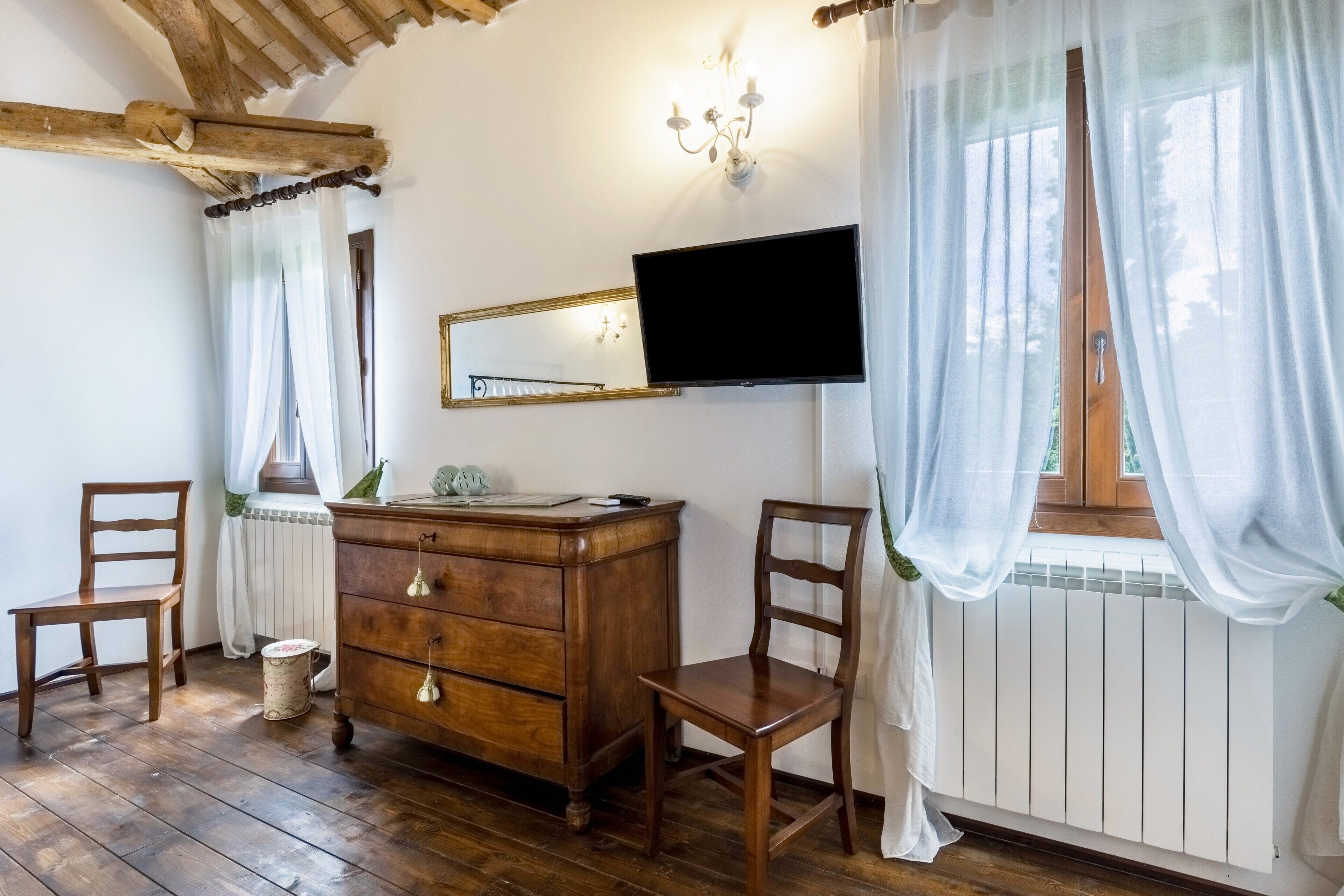Une pièce avec un plancher en bois, une commode en bois, deux chaises, un téléviseur fixé au mur et une fenêtre ornée de rideaux légers.