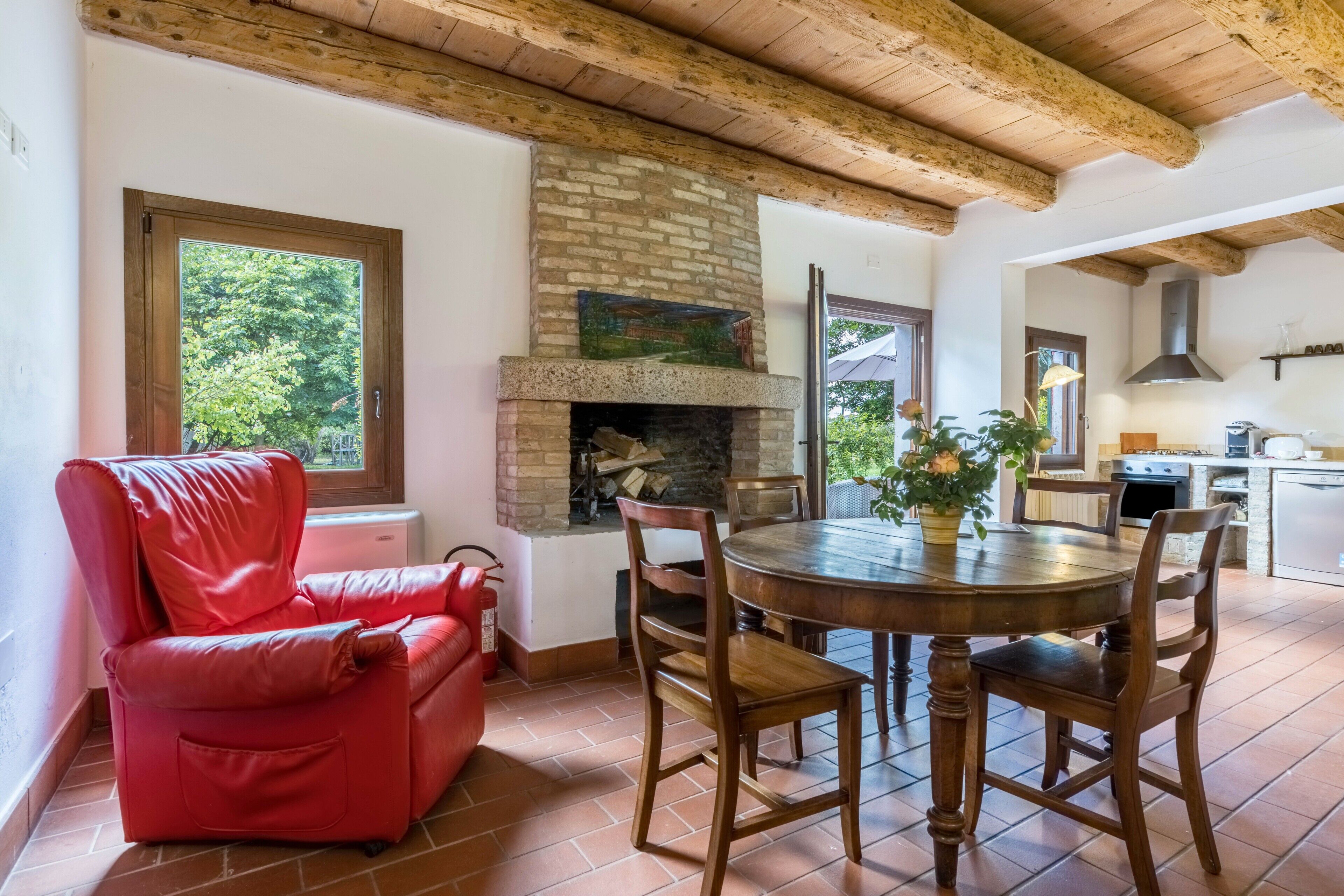 Une pièce avec une cheminée, une table à manger avec des chaises, un fauteuil en cuir rouge et une vue sur les arbres par la fenêtre.