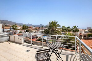 A rooftop terrace with a view of a coastal town, palm trees, and mountains in the distance.