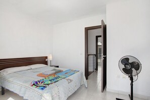 Une chambre à coucher avec un lit, un ventilateur sur pied et une porte donnant sur une autre pièce.