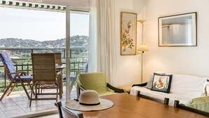 Un balcon donnant sur un paysage urbain, une table en bois, un fauteuil vert, un canapé blanc et un tableau encadré au mur.