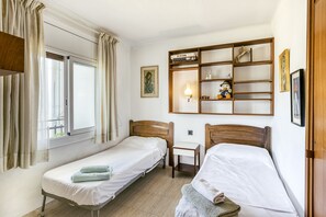 Een kamer met twee eenpersoonsbedden, een klein tafeltje, een plank met boeken en decoratieve objecten, en een raam met gordijnen.