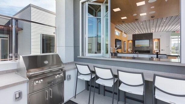 An outdoor kitchen area with a grill, bar seating, and a view of the interior through large windows. - Elan Menlo (Menlo Park)