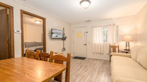 Quarto com mesa de jantar de madeira, cadeiras, sofá, televisão fixada na parede, porta com cortina e abajur sobre uma mesinha lateral.