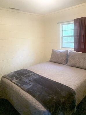 Una habitación pequeña con cama individual, paredes de ladrillo blanco, ventana con cortinas y ropa de cama estampada.