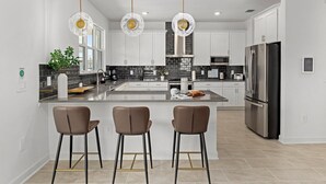 Una cocina moderna con armarios blancos, una isla grande con taburetes de bar, electrodomésticos de acero inoxidable e iluminación colgante.