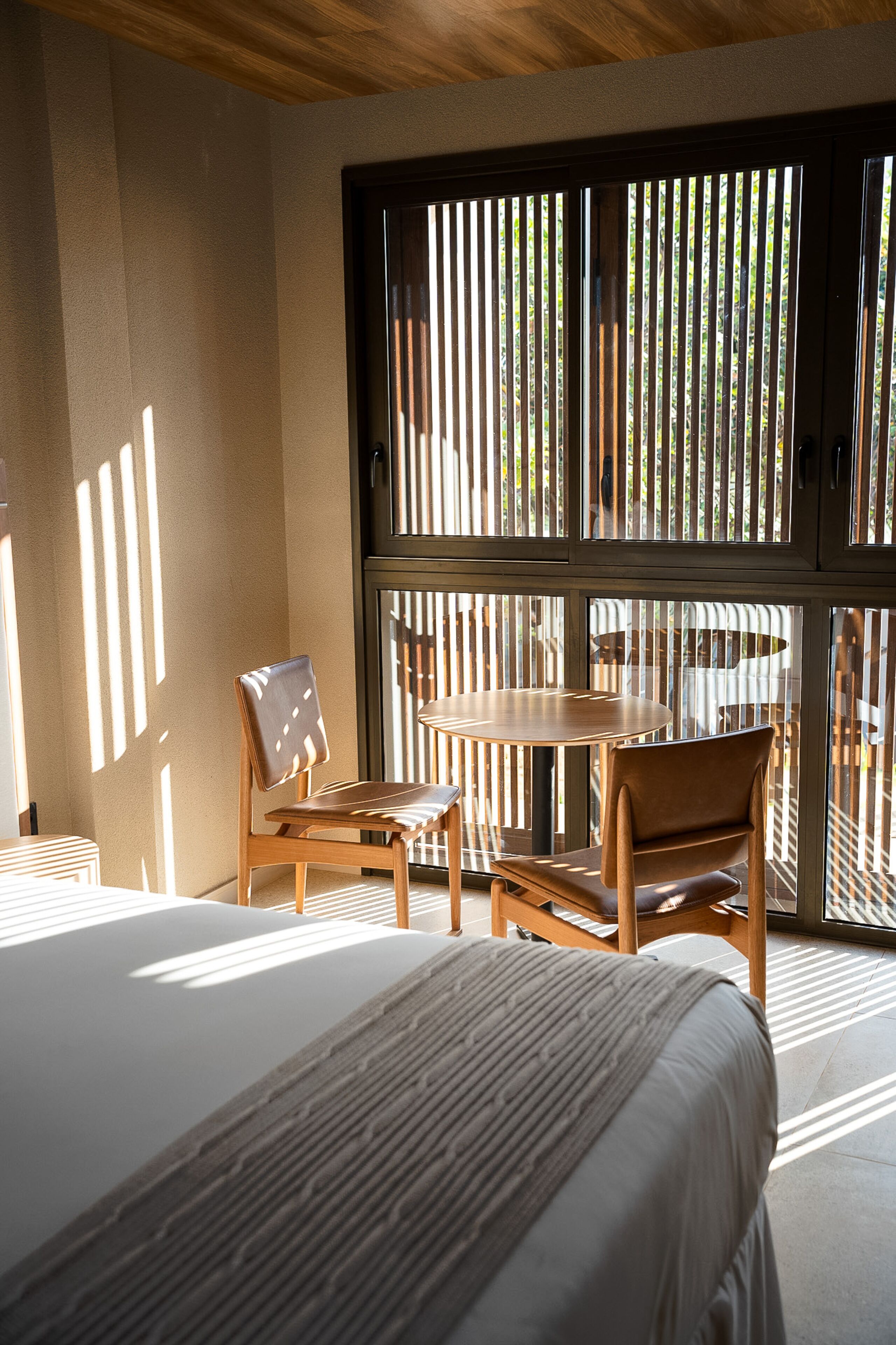 A bedroom with a large bed, a wooden chair, a small table, and a window with vertical blinds.