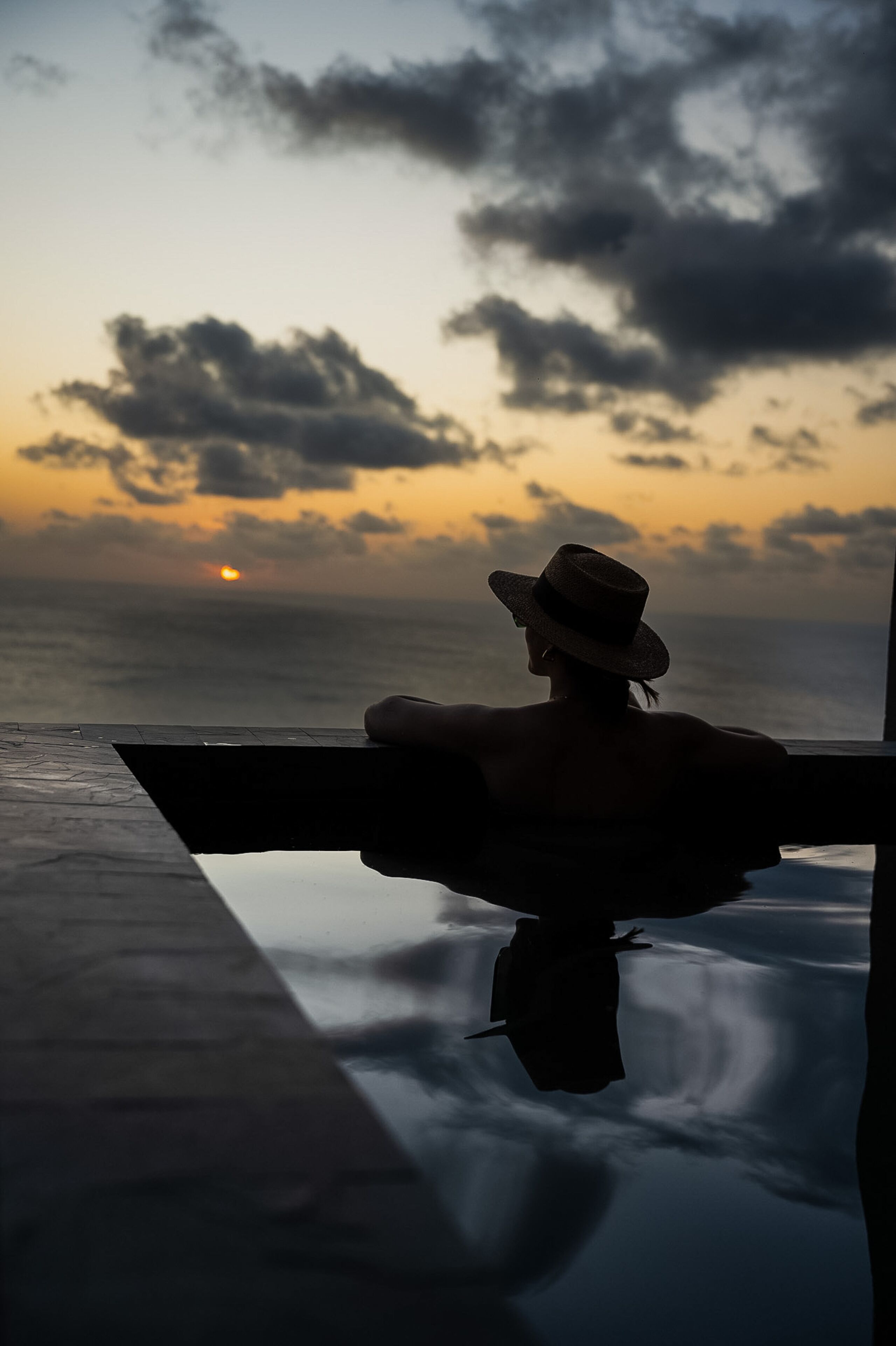A person in a hat sitting by a pool with a sunset view.