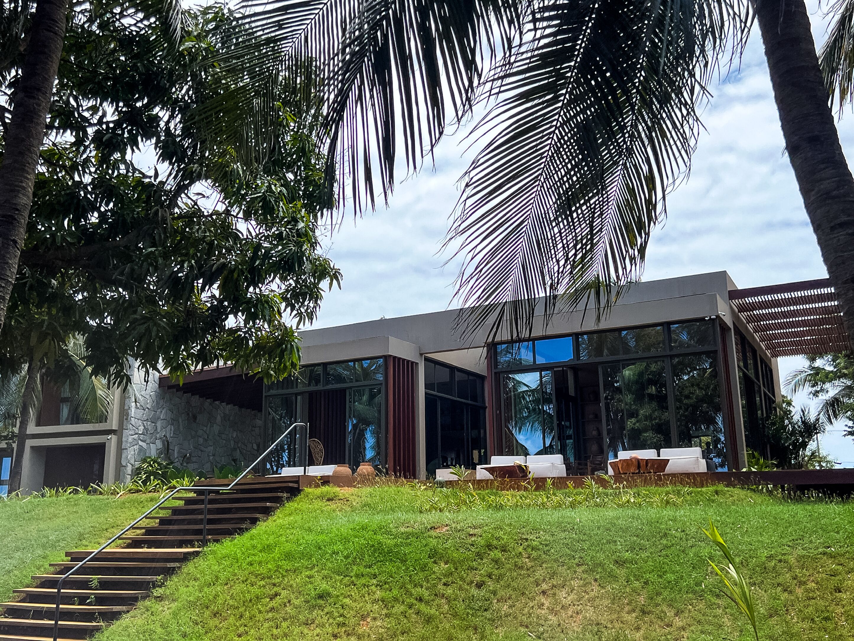 A modern house with a staircase leading to a glass door, surrounded by lush greenery and palm trees.