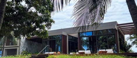 A modern house with a staircase leading to a glass door, surrounded by lush greenery and palm trees.