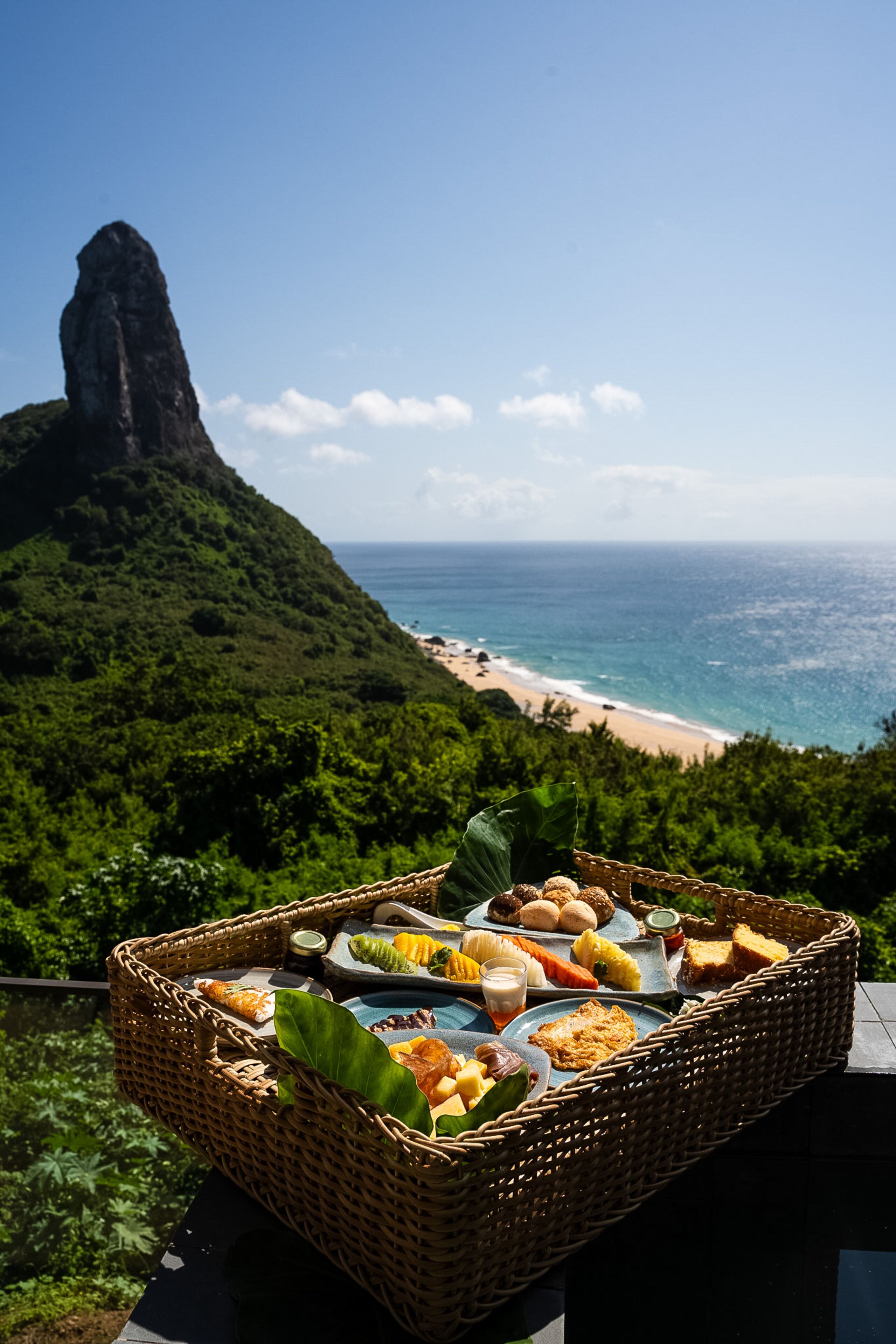 Un panier en osier rempli de nourriture et de boissons, posé sur un balcon donnant sur une plage et une montagne.