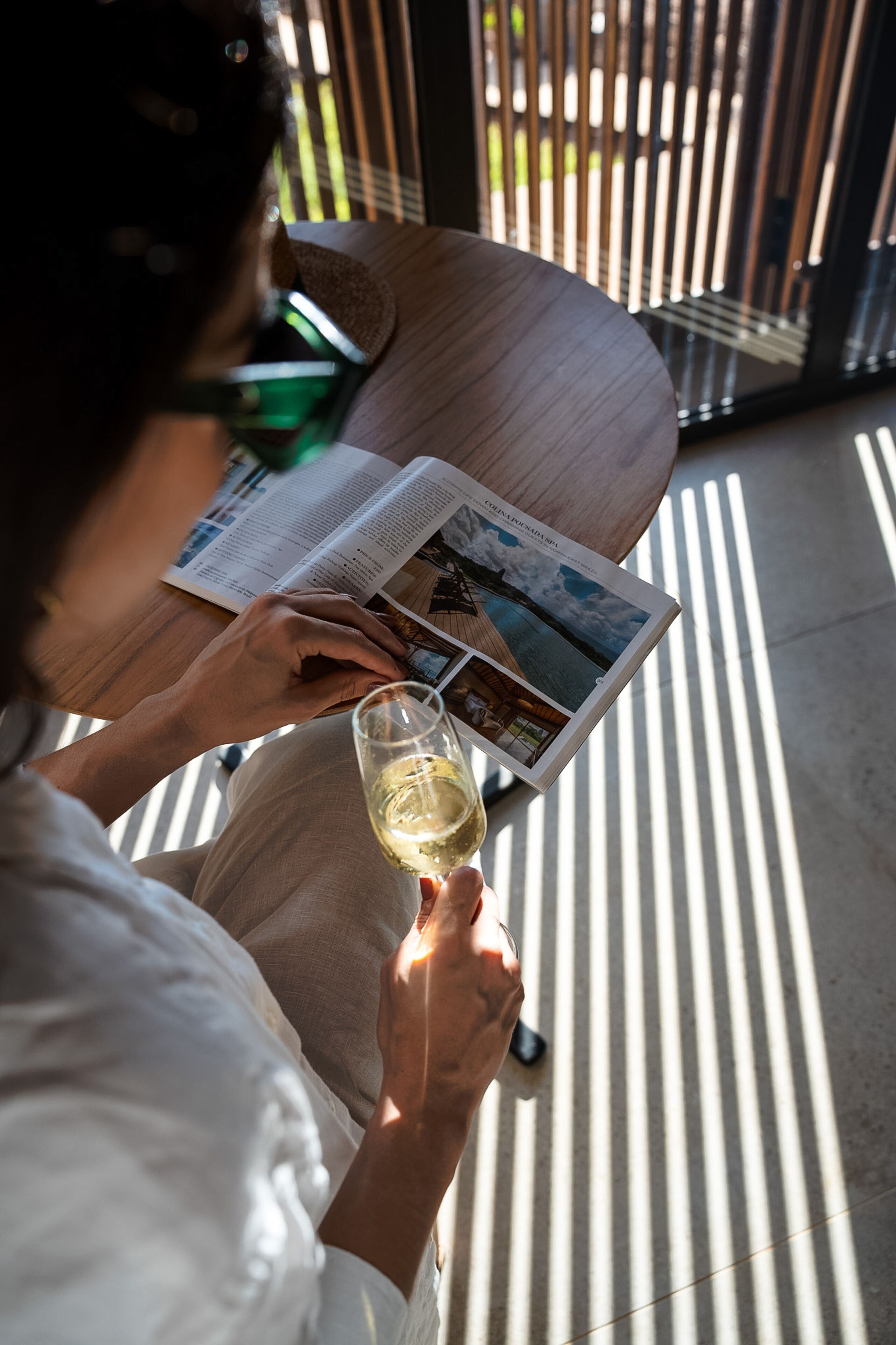 A person reading a magazine at a table with a glass of wine, near a window with vertical blinds.