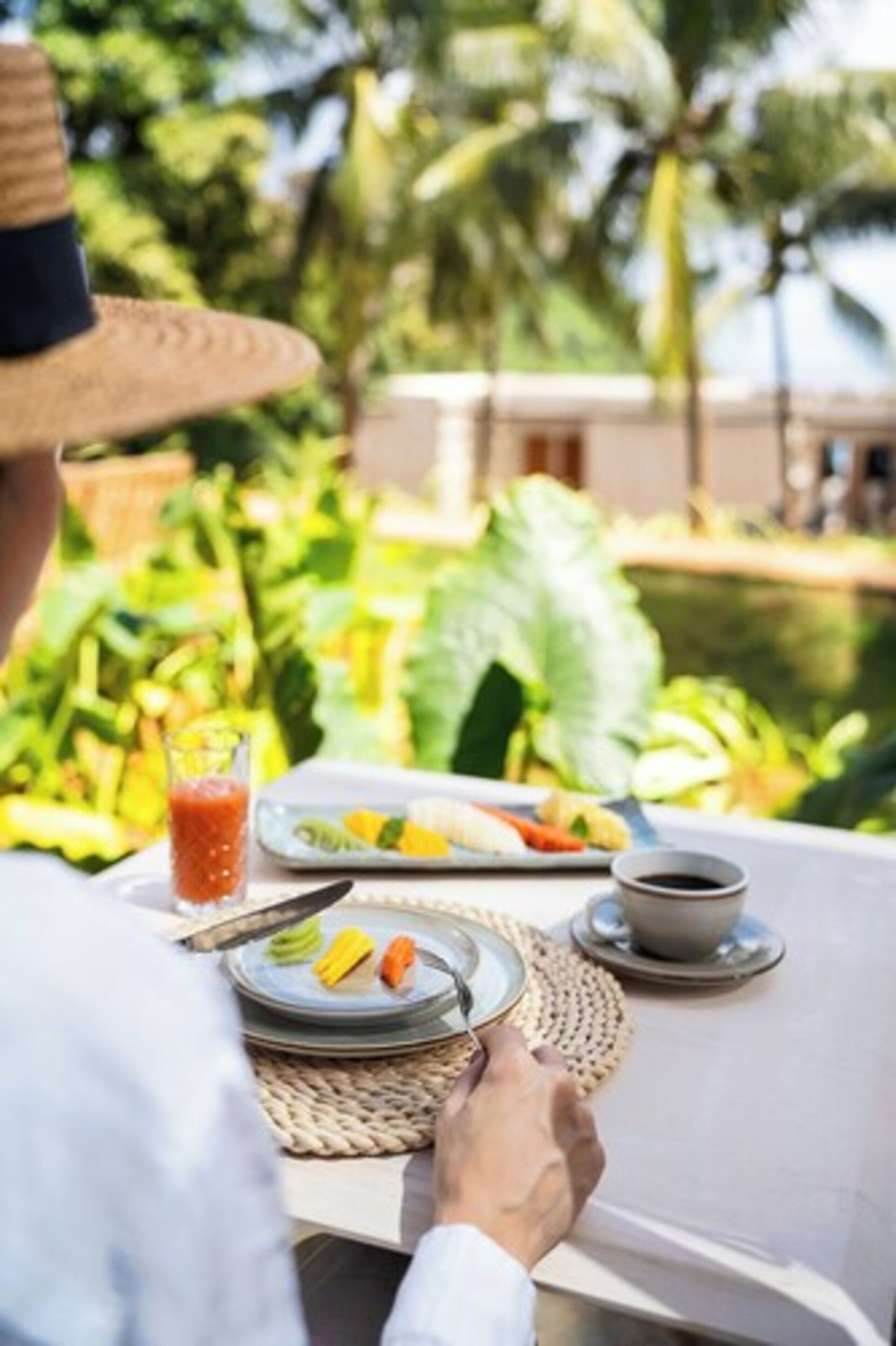 A dining table with plates of food, a glass of juice, and a cup of coffee, set against a backdrop of lush greenery and palm trees.