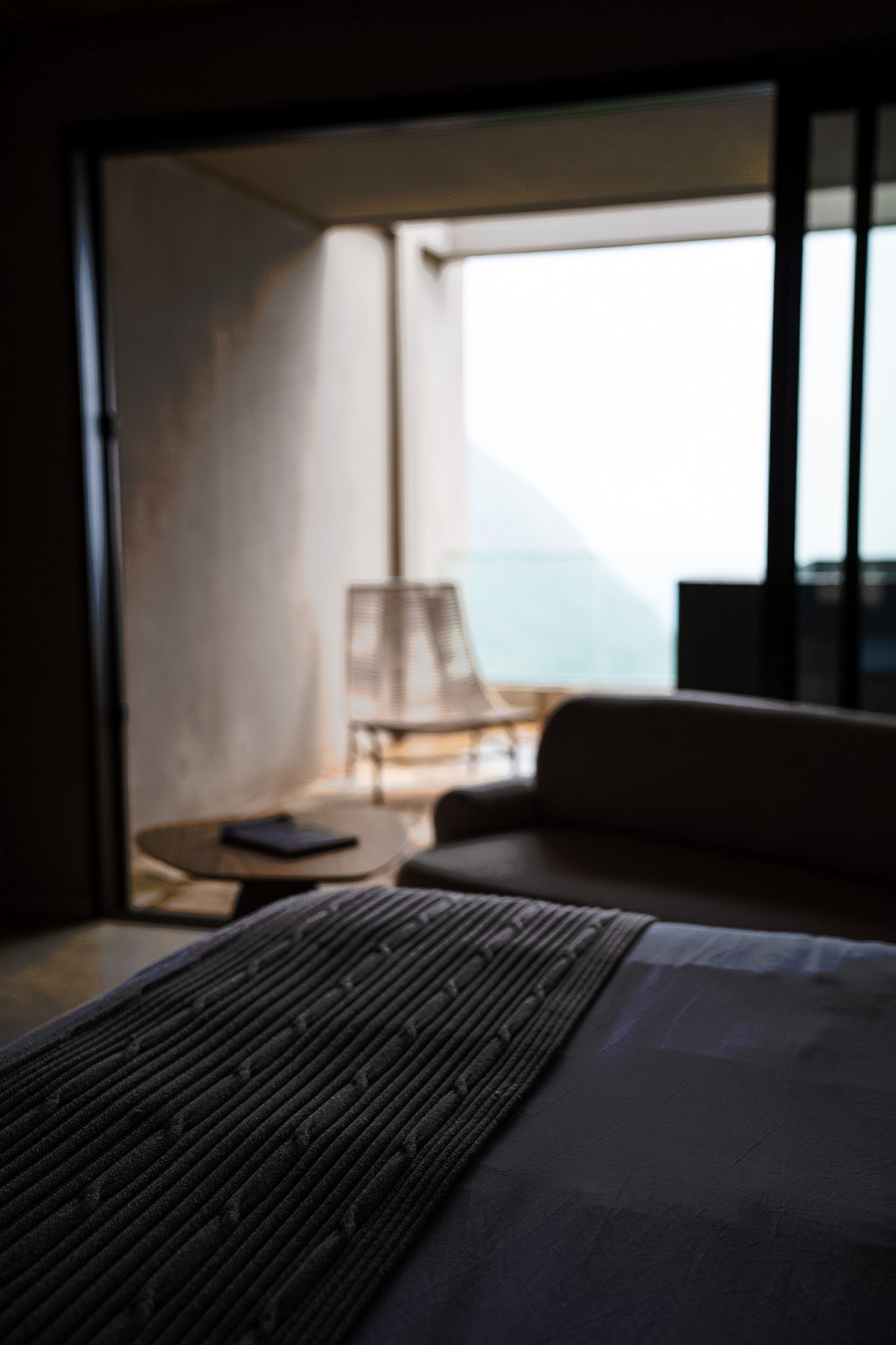 A hotel room with a bed, a small table, and a chair. There is a view of the ocean through large windows.