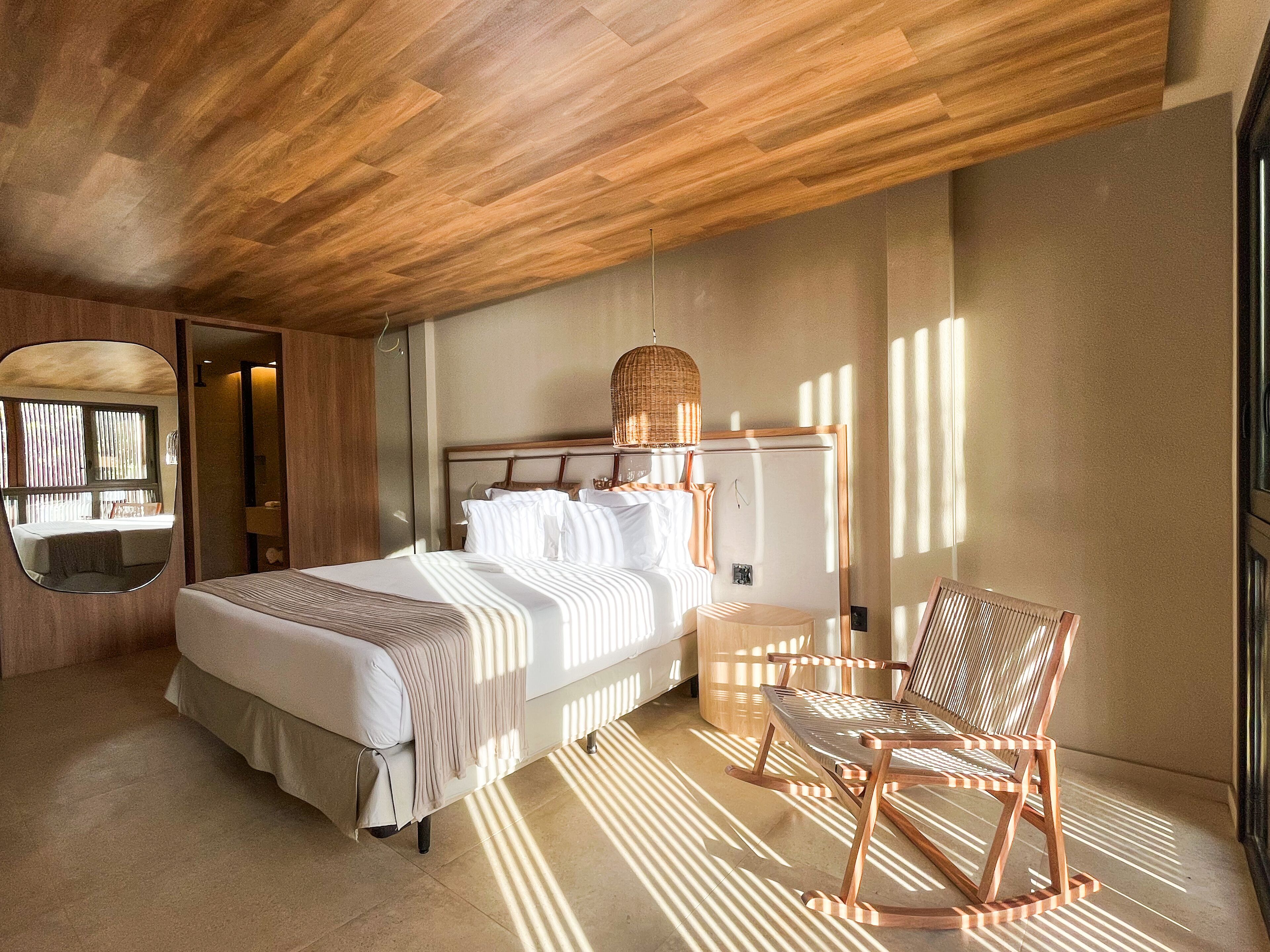A bedroom with a wooden ceiling, a bed with white linens, a wicker chair, and a large mirror on the wall.