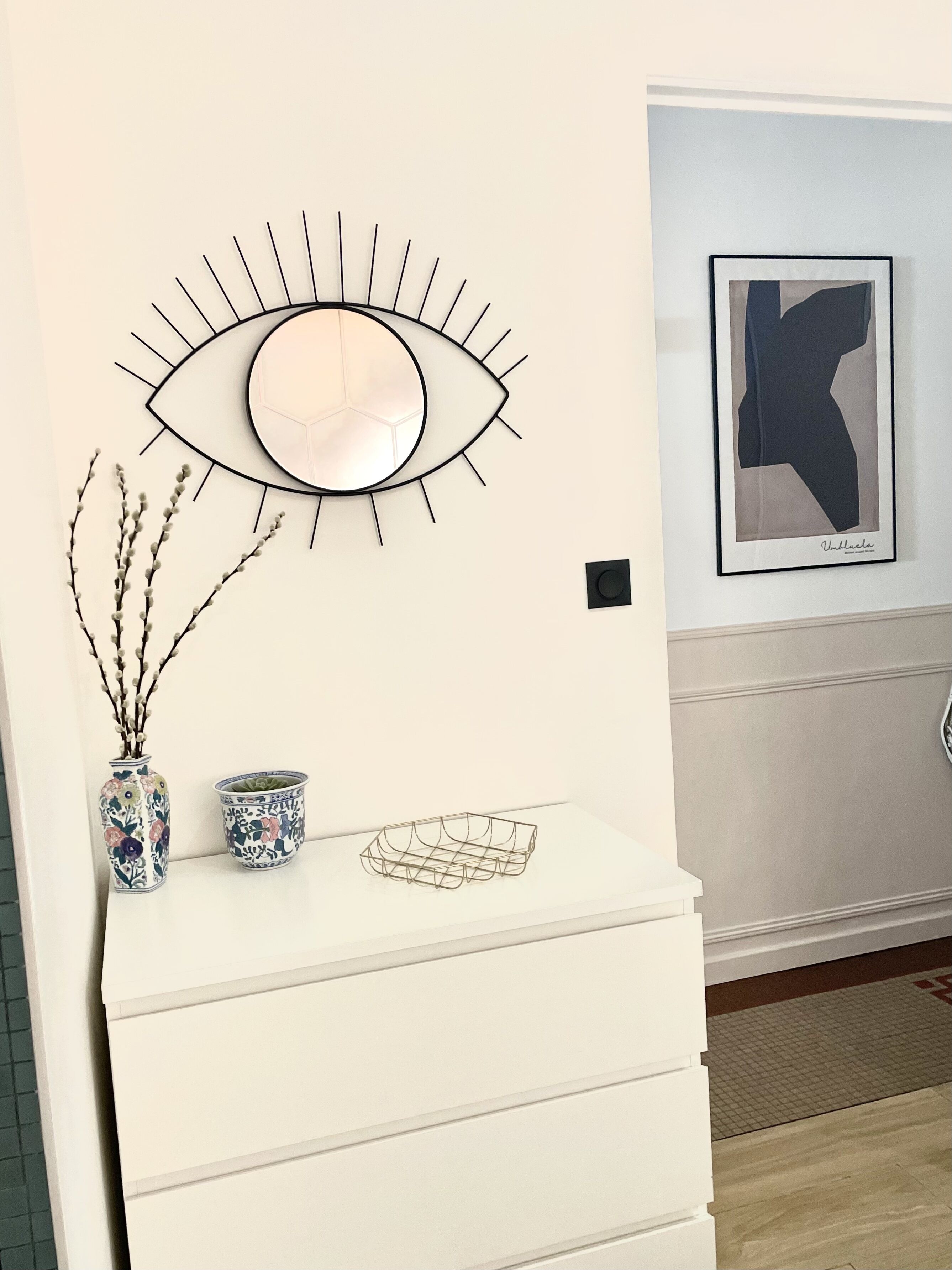 A white dresser with a decorative eye-shaped mirror above it, a framed abstract artwork on the wall, and two ceramic vases with floral patterns.