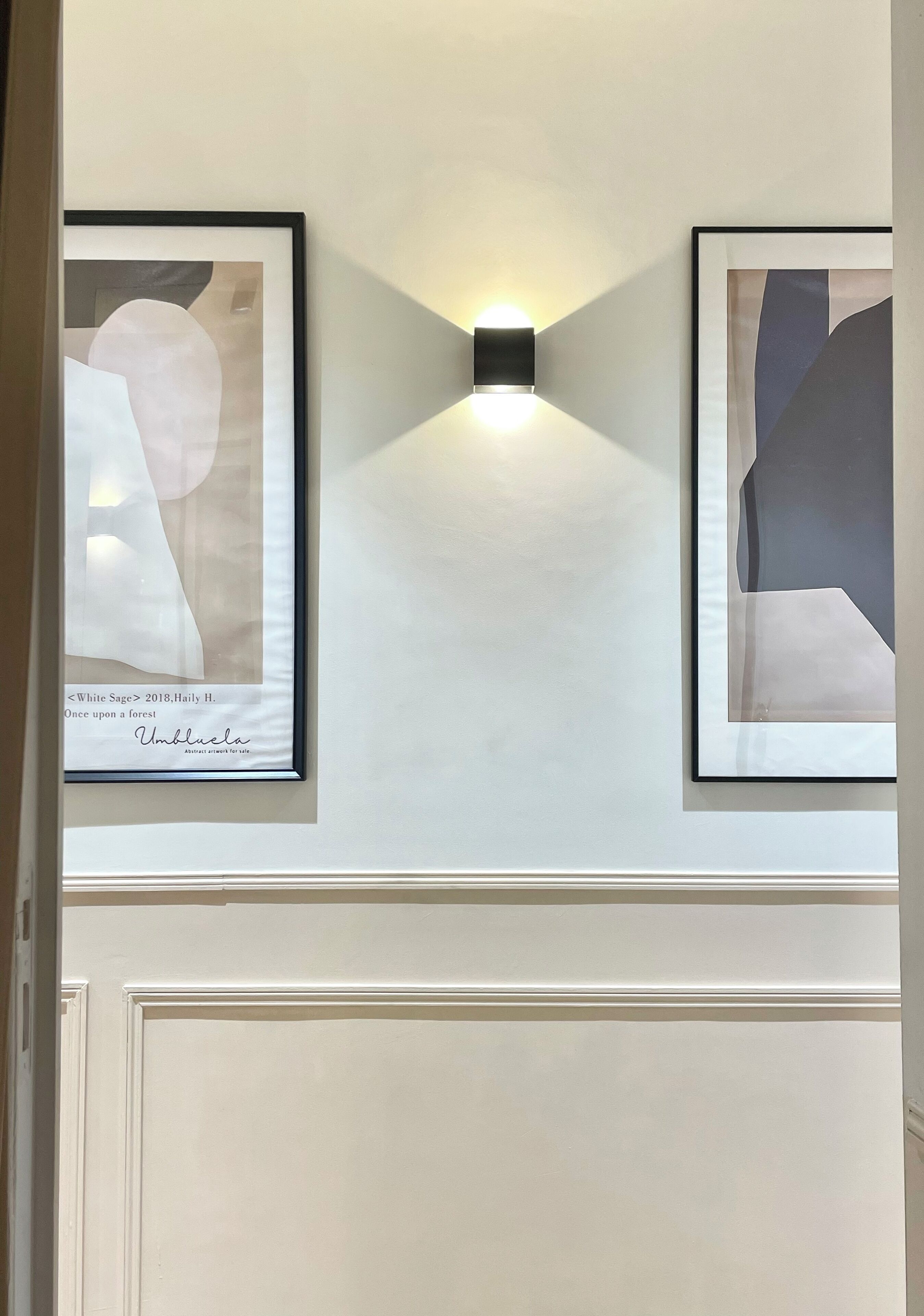 Dos cuadros abstractos enmarcados sobre una pared, con una pequeña luz de pared colocada entre ellos.