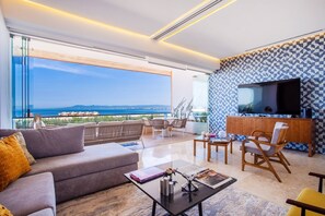 Una sala de estar moderna con sofá, mesa de centro y sillas, un televisor y un balcón con vistas al mar.