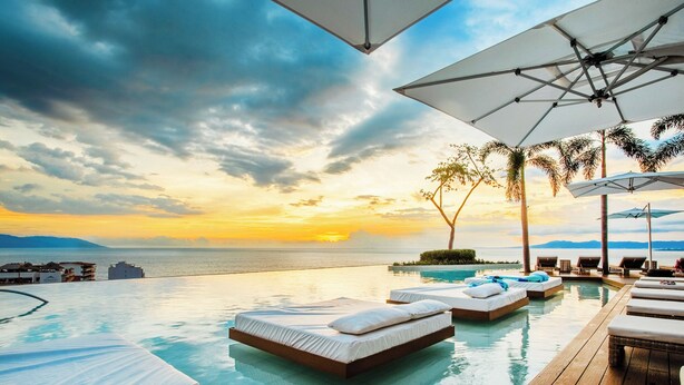 Une piscine à débordement avec des chaises longues et des parasols, offrant une vue imprenable sur le coucher de soleil sur la mer.