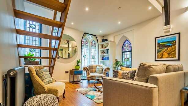 A cozy living room with a sofa, armchairs, and a wooden ladder. There are large windows with stained glass, a bookshelf, and a framed picture on the wall. - The Church Vaults (Matlock)