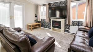 A living room with a brown leather sofa, a wooden coffee table, a flat-screen TV mounted above a fireplace, and patterned wallpaper. - Lake View Lodge (Windermere)