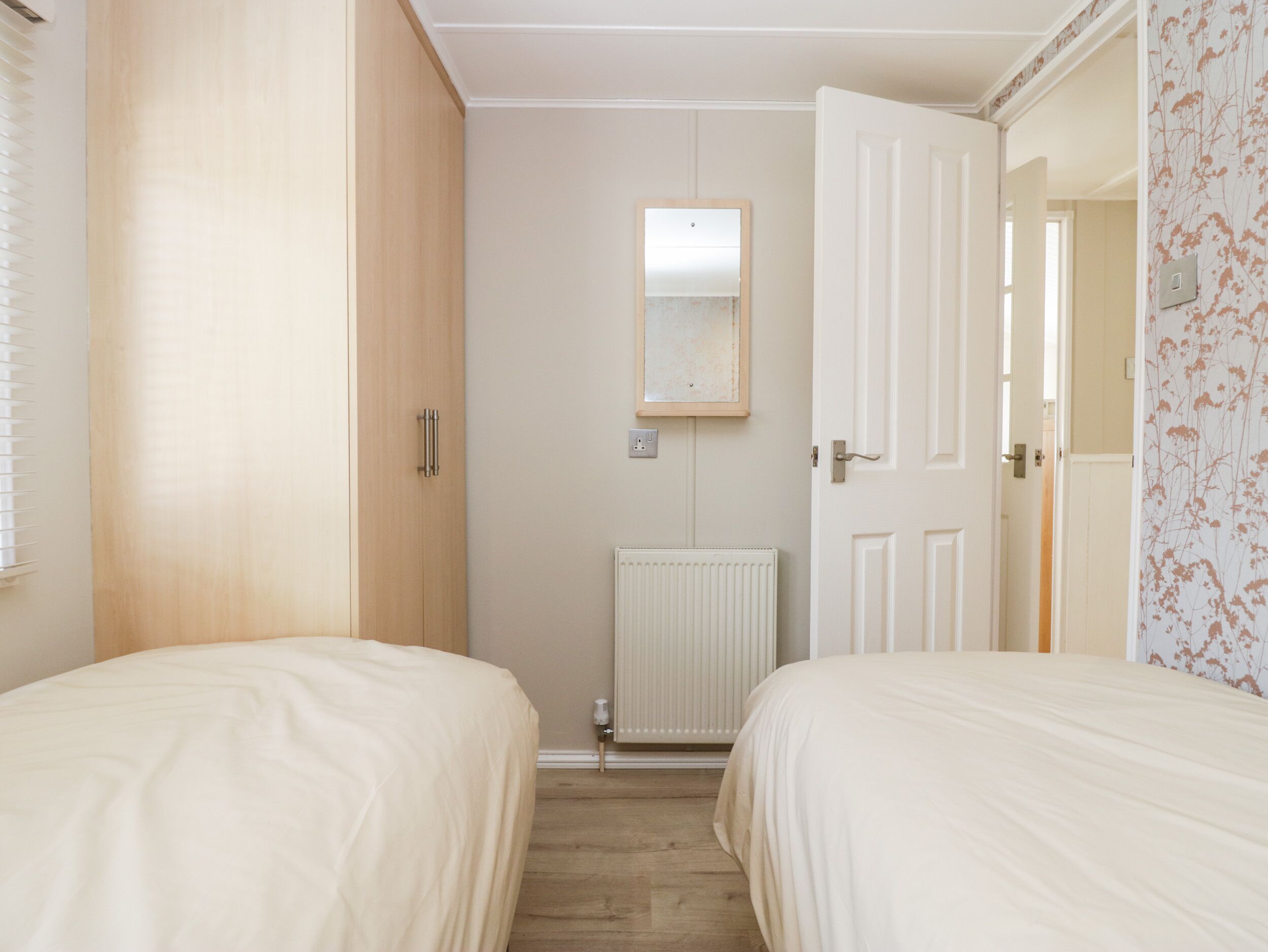 A bedroom with two beds, a radiator, a mirror, and a door leading to another room.
