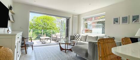 Ein geräumiges Wohnzimmer mit großem Fenster, einem grauen Sofa, Korbstühlen und einem Esstisch.